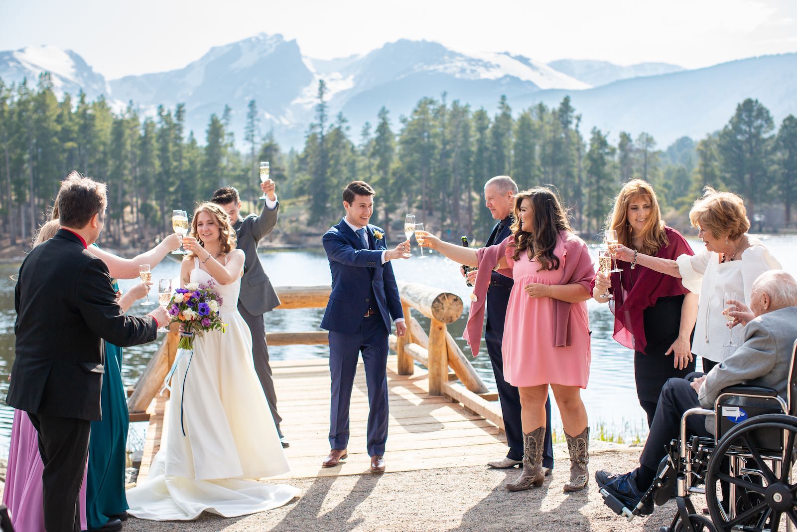 A wedding party raising champagne glasses on a dock at Sprague Lake in Rocky Mountain National Park